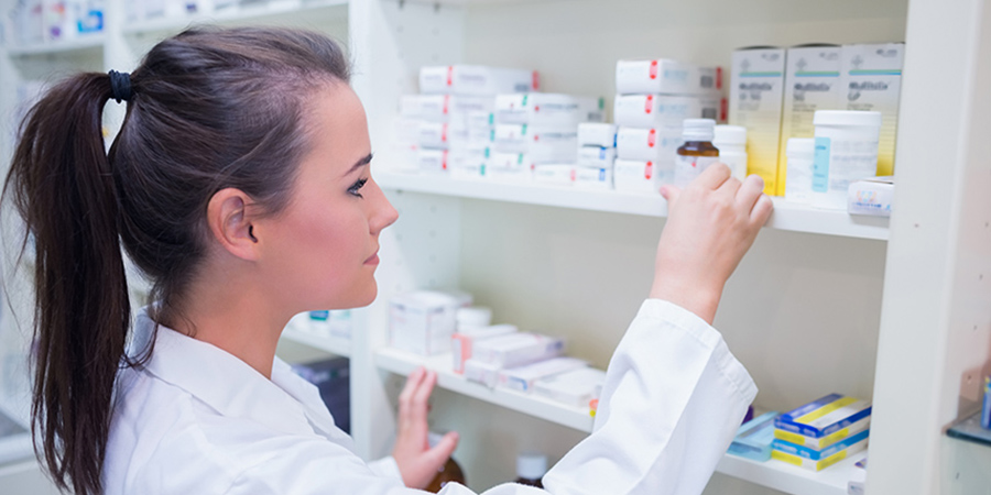 A young woman wearing a lab coat takes a bottle of medication from a pharmacy shelf