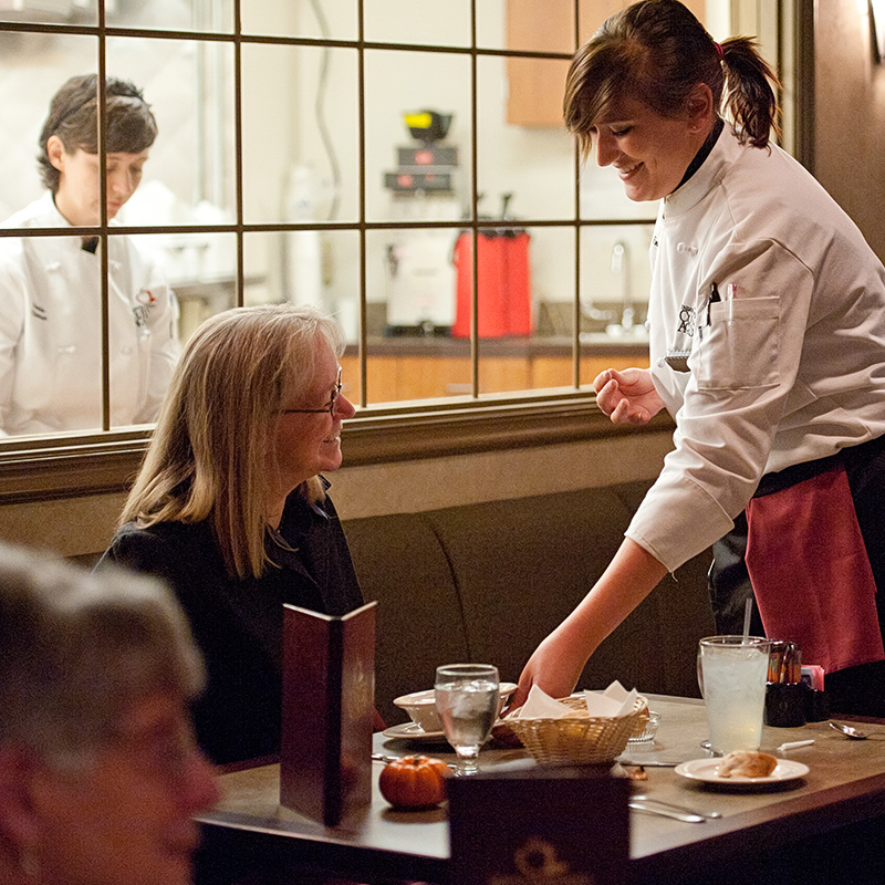 An SCC culinary student in a white chef garb serving food to a woman seated at a table.