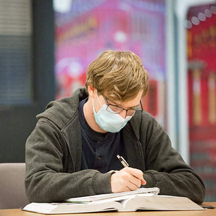 A male student seated at a table writing in his notebook. He