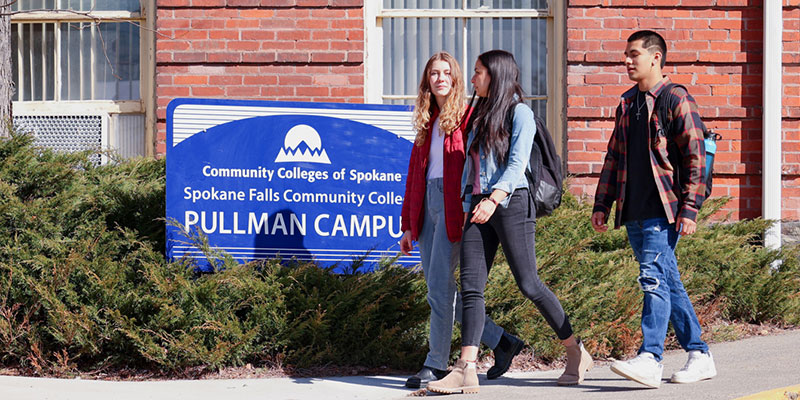Three students walking in front of the community colleges of Spokane Pullman Campus sign on a sunny day.