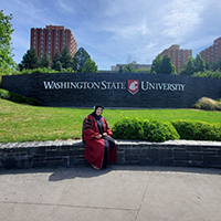 Dr. Intissar Yahia in PhD robes sitting outside in front of the Washington State University sign in Pullman.