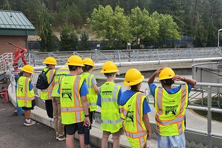 Eight elementary school children wearing hard hats and safety vests on a public works tour.