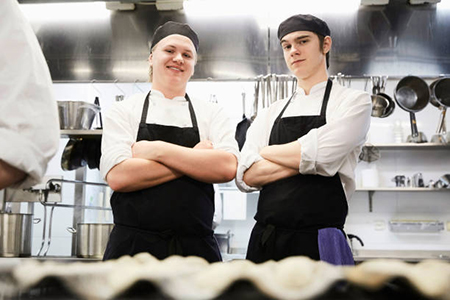 Two smiling chef students standing arms crossed in commercial-kitchen.
