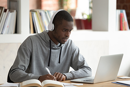 A person wearing headphones is sitting down at a table working on paper and is using a book and a laptop for reference material. 